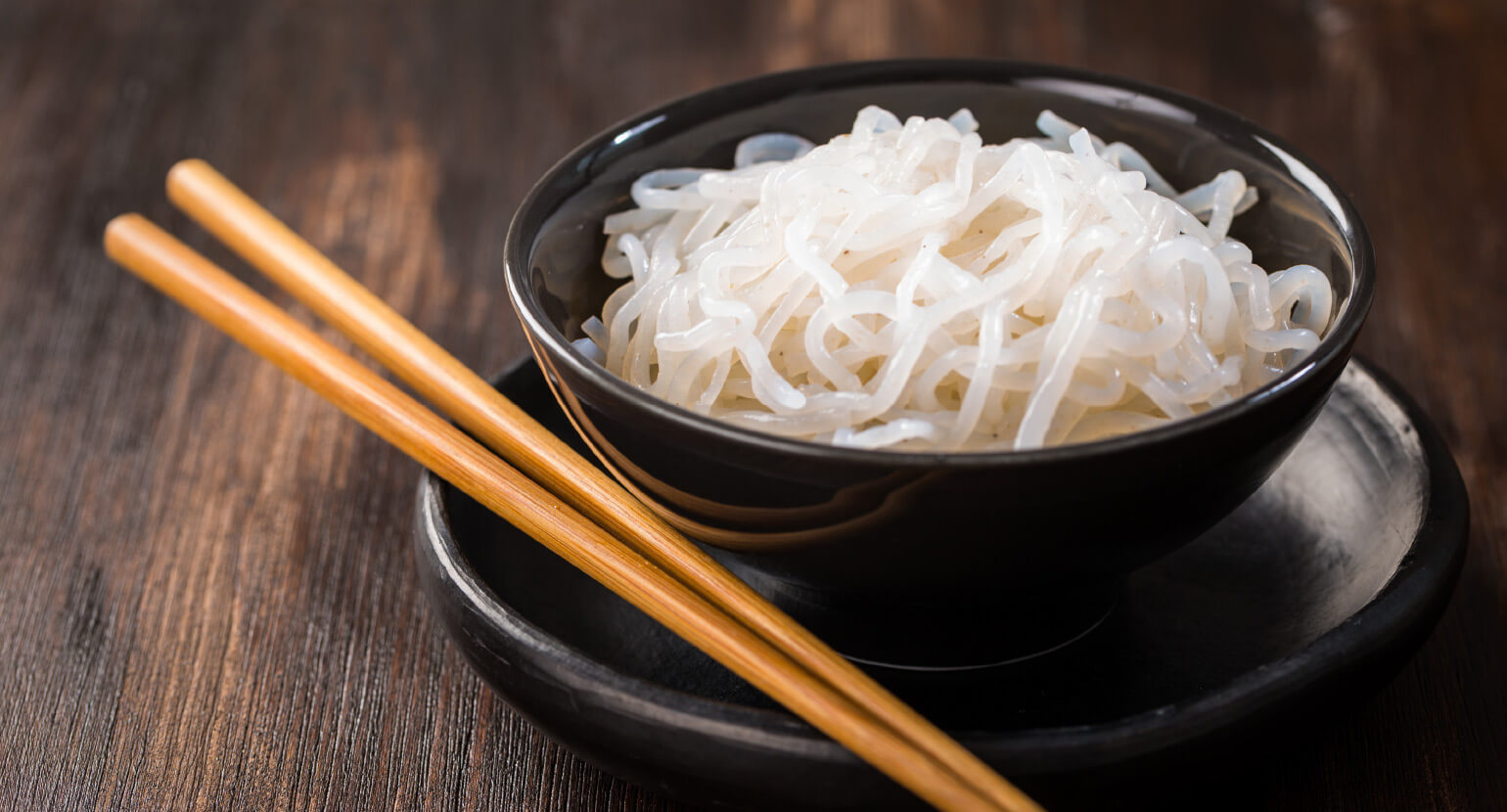 Natural Pharmaceuticals ReduXin Forte Juodos lėkštės ir dubenėlio su ryžių makaronais ir medinių lazdelių rinkinys ant tamsaus medinio stalo.